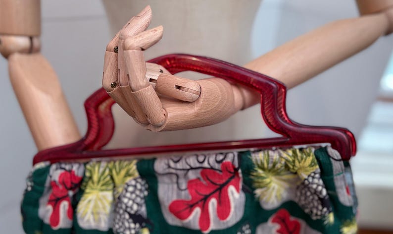 Peut inclure: Un sac &agrave; main vintage avec une poign&eacute;e rouge et un corps en tissu &agrave; motifs. Le tissu pr&eacute;sente des feuilles rouges, des ananas et d'autres motifs tropicaux sur un fond vert. La poign&eacute;e est en plastique rouge transparent incurv&eacute;.
