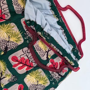 Peut inclure: Un sac fourre-tout vert avec un motif r&eacute;p&eacute;t&eacute; d'arbres, de feuilles et de pommes de pin rouges, jaunes et gris. Le sac a une poign&eacute;e bordeaux et une doublure int&eacute;rieure bleu clair. Le sac est en tissu.
