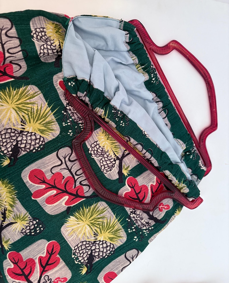 Peut inclure: Un sac fourre-tout vert avec un motif r&eacute;p&eacute;t&eacute; d'arbres, de feuilles et de pommes de pin rouges, jaunes et gris. Le sac a une poign&eacute;e bordeaux et une doublure int&eacute;rieure bleu clair. Le sac est en tissu.