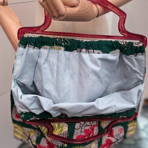 Peut inclure: Un sac &agrave; main vintage avec une poign&eacute;e rouge et un motif floral vert, gris et rouge. L'int&eacute;rieur est en tissu bleu clair. Le sac est tenu par une main de mannequin en bois.