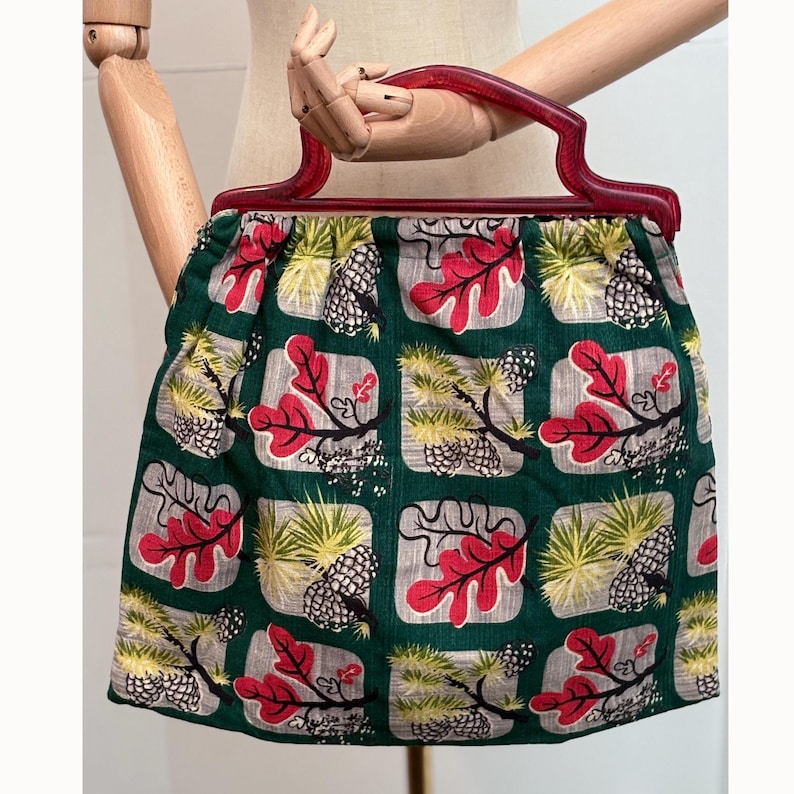 Peut inclure: Un sac &agrave; main de style vintage avec une poign&eacute;e rouge fonc&eacute; et un corps en tissu vert. Le tissu pr&eacute;sente un motif r&eacute;p&eacute;t&eacute; de feuilles rouges, de pommes de pin et de feuillage jaune-vert sur fond gris.