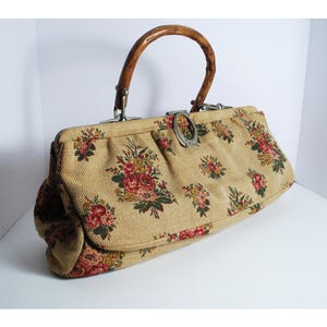 Peut inclure: Un sac à main vintage avec un extérieur en tissu beige orné de motifs floraux rouges, roses et verts. Il est doté d'une poignée en bois et d'un fermoir argenté. Le design du sac rappelle les styles vintage classiques.