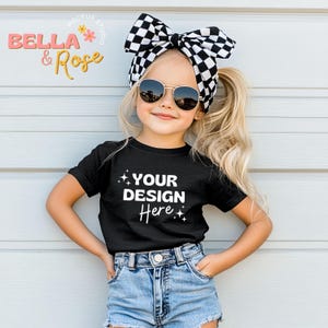Puede incluir: Una ni&ntilde;a peque&ntilde;a con una camiseta negra con el texto "YOUR DESIGN Here" en blanco, pantalones cortos de mezclilla y una diadema con lazo a cuadros en blanco y negro. Lleva gafas de sol. El fondo es una pared azul claro.