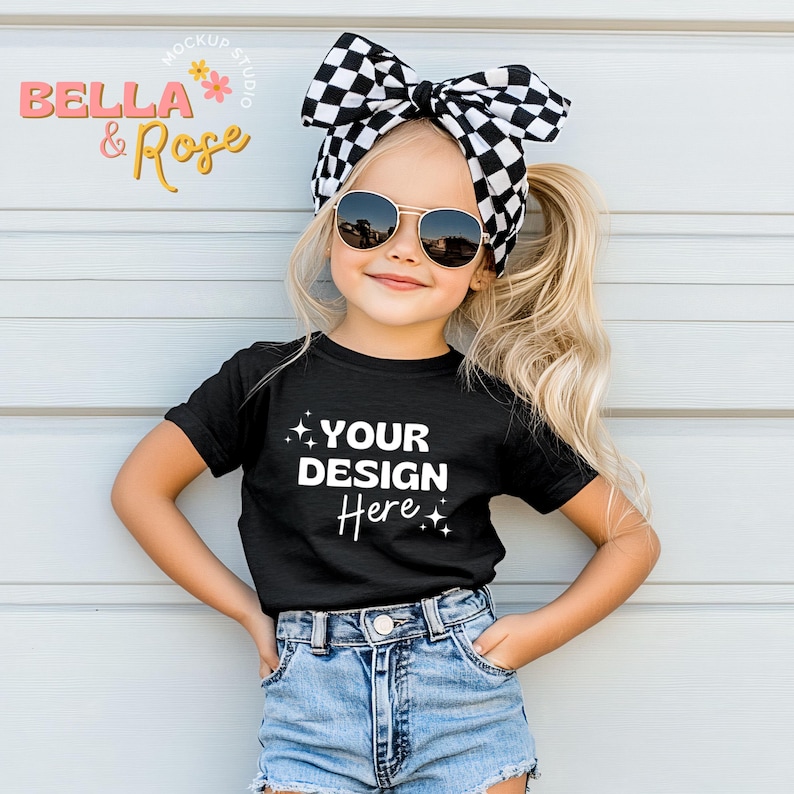 Puede incluir: Una ni&ntilde;a peque&ntilde;a con una camiseta negra con el texto "YOUR DESIGN Here" en blanco, pantalones cortos de mezclilla y una diadema con lazo a cuadros en blanco y negro. Lleva gafas de sol. El fondo es una pared azul claro.