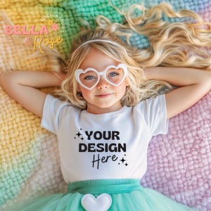 Puede incluir: Una niña pequeña con una camiseta blanca con el texto "YOUR DESIGN HERE" en ella, tumbada en una manta de colores arcoíris. Lleva gafas de sol en forma de corazón y una falda de tul verde.
