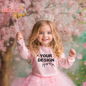 May include: A young girl wearing a pink sweatshirt with the text "YOUR DESIGN Here" and a pink tulle skirt. She is wearing a sparkly silver crown and holding a pink wand. The background is a pink and green floral backdrop.
