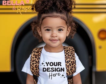 May include: A young girl wearing a white t-shirt with the text "YOUR DESIGN Here" in black. She is wearing a leopard print backpack and has her hair in two buns.