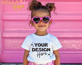 May include: A young girl wearing a white t-shirt with the text "YOUR DESIGN Here" and pink heart-shaped sunglasses. The girl has her hair in pigtails and is standing in front of a pink wall.