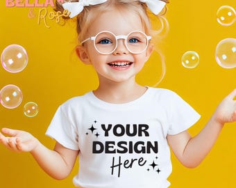 May include: A young girl wearing a white t-shirt with the text "YOUR DESIGN Here" in black. She is smiling and has her hands raised. The background is a bright yellow.