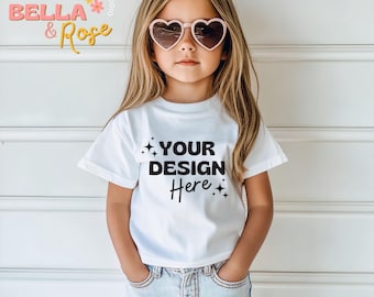 May include: A young girl wearing a white t-shirt with the text "YOUR DESIGN Here" and pink heart-shaped sunglasses. The girl is standing in front of a white wood panel background.