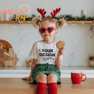 May include: A young child wearing heart-shaped sunglasses, red reindeer antlers, a white t-shirt with the text "YOUR DESIGN HERE", a green skirt, and red socks. The child is holding a cookie and sitting near a red mug.