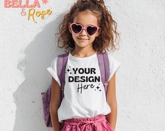 May include: A young person wearing a white t-shirt with the text "YOUR DESIGN Here" in black, paired with pink shorts and heart-shaped sunglasses. A purple backpack is worn over the shoulders. The background is a white wall.