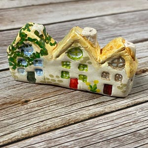 May include: A miniature ceramic house figurine with a thatched roof and painted details. The house features small windows, a chimney, and a door. The exterior is painted in cream, green, yellow, and red. The figurine is placed on a wooden surface.