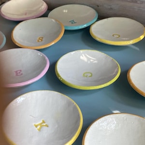 Könnte beinhalten: Eine Sammlung kleiner, runder, weißer Keramikschalen mit farbigen Rändern und einem einzelnen Buchstaben in der Mitte. Die Ränder und Buchstaben sind in verschiedenen Pastelltönen wie Gelb, Rosa, Orange und Hellblau gehalten.