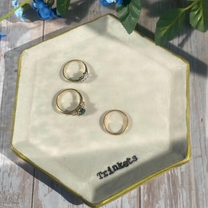 以下が含まれることがあります： 金色の縁が付いた六角形の小物入れに3つの指輪が置かれています。「Trinkets」の文字が黒で書かれた薄緑色の皿。背景には青い花と緑の葉があります。