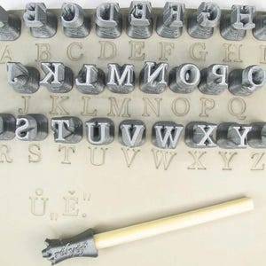May include: A set of gray letter stamps arranged on a beige surface, with corresponding letter imprints below. Includes a wooden handled stamp tool with the text "rélyef". Ideal for crafting and personalization projects.