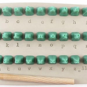 May include: A set of green alphabet stamps with a wooden handle. The letters a through z are arranged in rows, with the word "šňua" at the end. The stamps are on a beige background.
