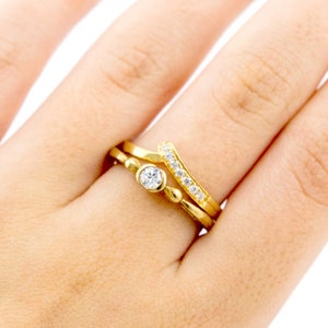May include: A gold ring set with a single round diamond, worn on a finger with two other gold bands. One band is plain and the other has a row of small diamonds.