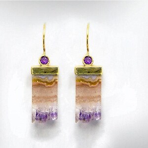 May include: A pair of gold-tone earrings featuring rectangular, layered gemstone pendants with shades of purple, tan, and white. Each earring has a small, round purple gemstone and a gold-tone hook.