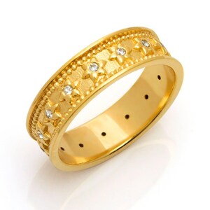 May include: A gold-colored ring with a floral pattern and small, clear gemstones. The ring has a textured band with a repeating star-like design and a smooth, polished interior. The ring is set against a white background.