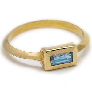 May include: A gold ring featuring a rectangular, light blue gemstone. The ring has a simple band and a bezel setting. The ring is displayed against a white backdrop.