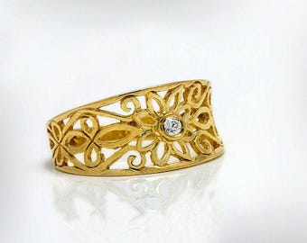 May include: A gold-colored ring with an openwork floral design. The ring features a central flower with a small, clear gemstone. The band has a wide, curved shape with intricate cut-out patterns.