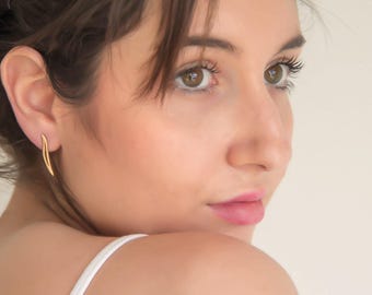May include: A close-up shot of a woman wearing a pair of gold earrings. The earrings have a curved, elongated design. The woman has brown hair and is looking over her shoulder. She is wearing a white top.