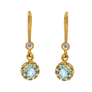May include: A pair of gold-coloured drop earrings. Each earring features a round, light blue gemstone set within a decorative gold filigree frame. A small, clear gemstone is set above the blue stone. The earrings have a textured, ornate design.