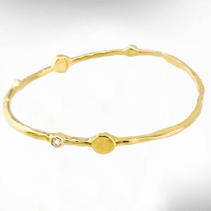 May include: A gold-coloured bangle bracelet with a textured, organic design. The bracelet features a small, round accent and a tiny, sparkling gemstone. The overall aesthetic is minimalist and elegant, suitable for everyday wear.