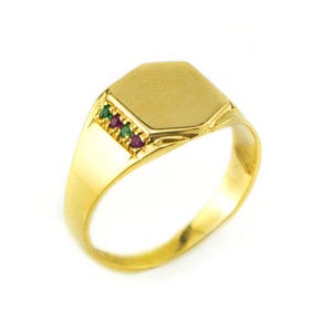 Op de afbeelding: Een gouden zegelring met een rechthoekige bovenkant en een band versierd met kleine groene en rode edelstenen. De ring is gemaakt van gepolijst goud tegen een witte achtergrond.
