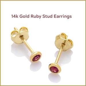 May include: A pair of 14k gold stud earrings featuring ruby gemstones. The earrings have a post and backing design. The ruby gemstones are oval-shaped and set in a bezel setting. The text "14k Gold Ruby Stud Earrings" is displayed above the earrings.