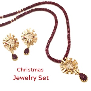 Könnte beinhalten: Ein goldfarbenes Schmuckset mit Halskette und Ohrringen mit floralem Design. Die Halskette und die Ohrringe haben einen tiefroten Edelstein. Das Set befindet sich auf weißem Hintergrund. Der Text "Christmas Jewelry Set" ist rot.