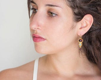 May include: Gold-toned dangle earrings with a red oval stone, a small turquoise stone, and a dangling red bead. The earrings have intricate filigree detailing and a hook closure. The earrings are worn on a person.