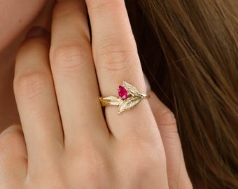 Könnte beinhalten: Ein goldfarbener Ring mit Blattmotiv und einem tropfenförmigen, roten Edelstein. Der Ring hat ein detailliertes Band und Blätter. Der Ring wird an einem Finger getragen. Das Design erinnert an die Natur.