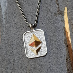 May include: Silver pendant necklace with a rectangular pendant featuring a textured background and two gold-colored geometric shapes. The necklace has a silver and black rope chain.