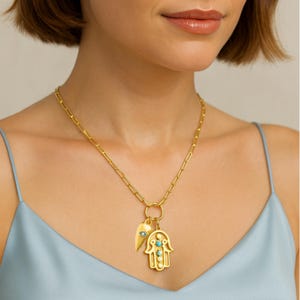 May include: Gold-coloured necklace featuring a paperclip chain and two pendants. One pendant is a Hamsa hand with turquoise details, and the other is a gold leaf with a turquoise eye. The necklace is worn on a person wearing a light blue top.
