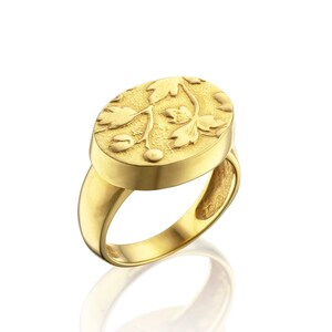 May include: A gold ring with an oval face featuring a detailed floral design. The ring's band and face are a bright, polished gold, reflecting light. The floral design includes leaves and small berries, creating a nature-inspired aesthetic.
