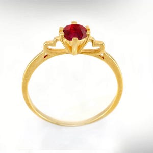 Può includere: Un anello d'oro con una gemma rotonda di colore rosso intenso. L'anello presenta un design delicato con accenti a forma di cuore sull'incastonatura. La fascia è liscia e lucida, creando un look classico ed elegante. L'anello è su sfondo bianco.