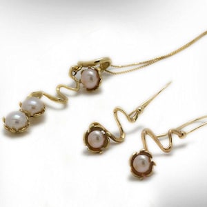 May include: A gold-toned jewelry set featuring a necklace with a pendant and a pair of earrings. The pendant and earrings have a wavy design with a pearl at the center of each piece. The necklace has a delicate chain.