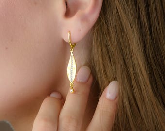 Può includere: Orecchini pendenti color oro. Ogni orecchino presenta un pendente a forma di goccia con una fila di piccoli cristalli trasparenti al centro. Un piccolo cristallo rotondo pende nella parte inferiore del pendente. Gli orecchini sono attaccati a un cerchio d'oro.