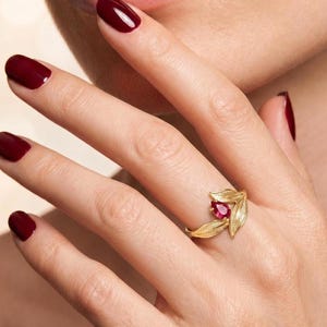 May include: A gold ring featuring a leaf design with a teardrop-shaped red gemstone. The ring is worn on a hand with polished burgundy nails. The ring's intricate details and the gemstone's color create a striking contrast.
