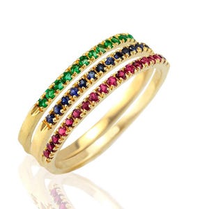 May include: Three gold rings, each adorned with a row of gemstones. One ring features green stones, another blue, and the third red. The rings are displayed on a white surface, reflecting the light.