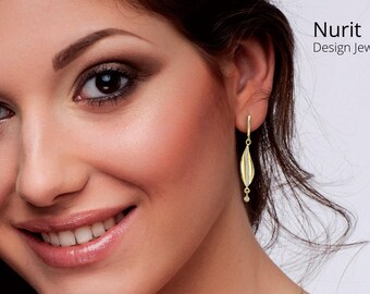 Può includere: Primo piano di una donna con un orecchino d'oro. L'orecchino ha un design a forma di foglia con un piccolo accento pendente. Il testo "Nurit Design Jewelry" è in alto a destra.