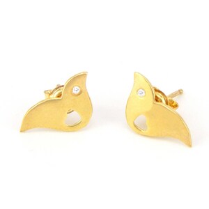 May include: A pair of gold-colored stud earrings. Each earring has a stylized bird or leaf shape with a small, round, clear gemstone and a cutout. The earrings are displayed on a white background.