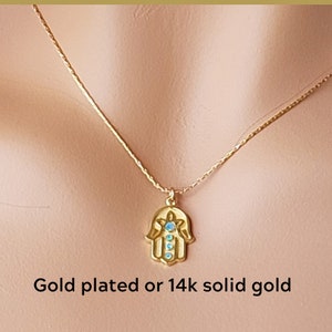 Puede incluir: Un collar de cadena de oro con un colgante de mano de hamsa con piedras azules. El collar está disponible en chapado en oro o en oro sólido de 14 quilates.