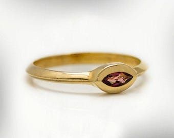 May include: A gold ring with a marquise-cut, reddish-pink gemstone. The ring has a smooth, polished band and a bezel setting that holds the gemstone securely. The ring is set against a white background.