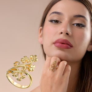 May include: A gold-colored ring with a floral design. The ring features a curved band and an ornate design of flowers and vines. The ring is shown on a person's finger, showcasing its intricate details and elegant style.