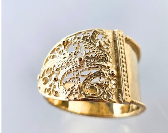 Könnte beinhalten: Ein goldfarbener Ring mit einem aufwendigen filigranen Design. Der Ring hat ein rechteckiges Band und eine gewölbte Oberseite mit einem detaillierten, spitzenartigen Muster. Das Metall hat eine polierte, glänzende Oberfläche.