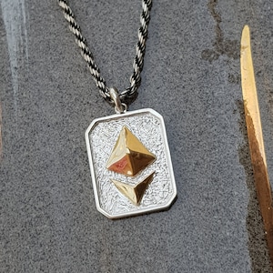 May include: A silver chain necklace with a silver and gold pendant featuring the Ethereum cryptocurrency symbol.
