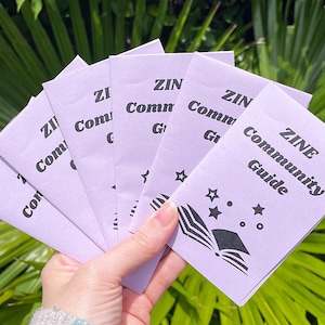May include: A hand holds a fan of lavender-colored ZINE Community Guides. Each guide features the title in black text, along with a graphic of an open book, stars, and dots. The background is a lush green foliage.